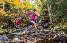 Man And Woman Trail Runners Rock-hopping In A River With Fall Foliage