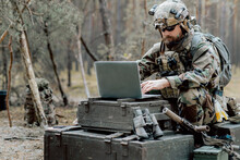 Bearded Soldier In Uniform Sit On Military Transport Crates, Analyze Data On A Laptop And Work Out Tactics At A Temporary Forest Base. In The Background, You Can See A Soldier Protecting The Base.