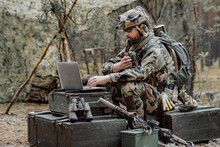 Bearded Soldier In Uniform Sit On Military Transport Crates, Analyze Data On A Laptop And Work Out Tactics At A Temporary Forest Base. In The Background, You Can See A Soldier Protecting The Base.