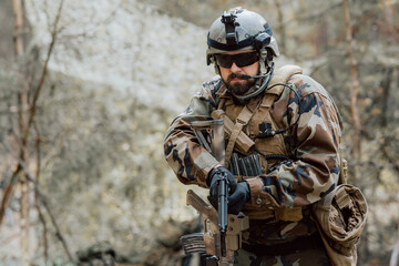 Portrait of a middle-aged bearded soldier in a Woodland military uniform and helmet, with headphones on his head holding a gun.