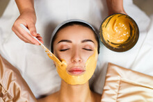 Cosmetic Jelly Mask, Facial Skincare. Cosmetologist Applies Yellow Gold Alginate Mask With Spatula On Face Of A Woman. Facial Skin Treatment In The Beauty Salon. Top View Above