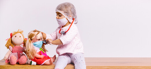 Banner,long format. A cute girl plays a doctor in a white coat, in a mask and with a stethoscope treats dolls. White background and side advertising space.