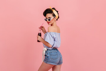 Sticker - Slim pinup girl in denim shorts looking over shoulder. Studio shot of beautiful young woman with bottle of soda standing on pink background.