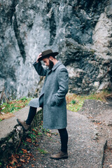 Stylish young man with a beard. He is wearing a coat and a hat on his head. During rainy weather in the mountains.