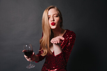 Wall Mural - Stylish lady with wineglass sending air kiss. Studio shot of blonde girl in red dress drinking wine at party.