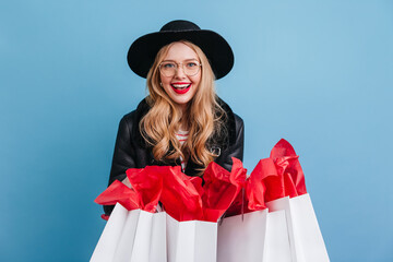 Wall Mural - Jocund blonde woman in hat holding shopping bags. Cheerful curly girl laughing on blue background.