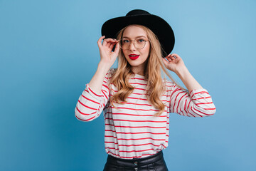 Wall Mural - Curly woman in elegant hat looking at camera. Studio shot of fascinating blonde girl in striped shirt.