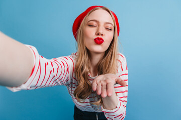 Wall Mural - Romantic blonde girl sending air kiss. Studio shot of french lady taking selfie on blue background.