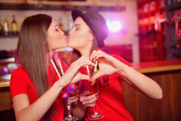 Two young lesbian girls kiss and make a heart with their hands at a club party