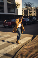 Wall Mural - Stylish handsome African male in a coat crossing the road and talking by phone