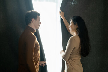 Poster - Young man and his wife looking at one another while drawing dark curtains