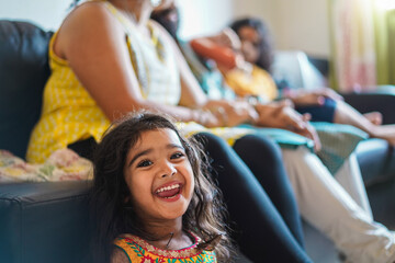 Wall Mural - happy indian girl child wearing sari dress sitting on sofa with parents at home - focus on girl face