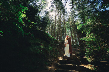 Wall Mural - Happy stylish couple newlyweds in the green forest on summer day. bride in long white dress and groom in red suit are hugging. wedding day.