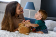 © lordn - Smiling mother playing with her cute toddler son at home in the bed. Using plush bear and baby toys. Maternity concept. Playing with children.