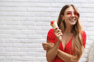 Happy blond girl travelling around city on summer day, eating sweet ice cream and smiling, trying local food on vacation. Young woman in sunglasses and dress standing happy near white wall outdoors