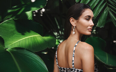 Poster - Tropical vacation and travelling concept. Young brunette woman with tanned skin, wearing bikini on tropic nature, standing near palm jungle leaves and looking aside