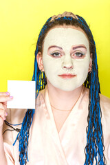 Woman with blue afro braids face in a frozen mask of green clay with a business card in her hands on a yellow background.