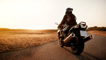 Handsome Motorcyclist In Leather Jacket And Helmet At Sunset On The Road In Warm Sun Rays
