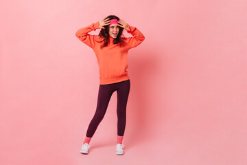 Wall Mural - Snapshot in full growth of young dark-haired girl in bright sports suit in style of 80s. Woman in pink sports headband posing on isolated background