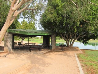 A covered seating area in a park with a picnic table and a man-made pond in the background in Arizona on a hot summer day