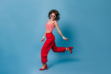 Sticker - Fashionable girl in clothes in style of 80s runs on blue background. Brunette in stylish glasses smiling