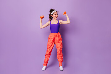 Wall Mural - Brunette woman in cap is engaged in fitness on purple background. Girl is in great mood holding red dumbbells