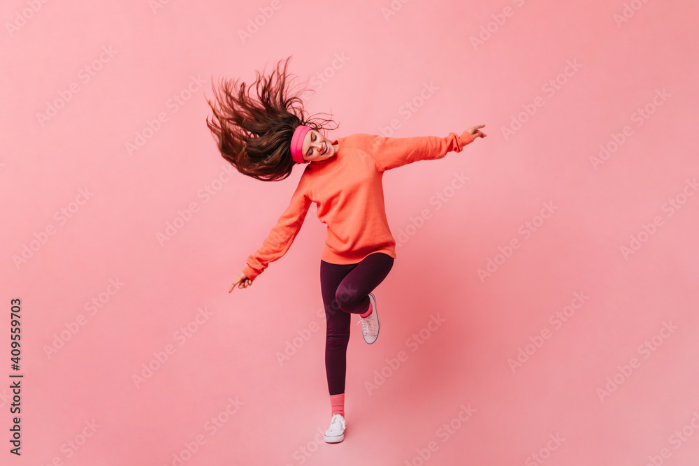 Young lady in sports outfit dancing on pink background. Full length portrait of woman in orange sweatshirt - obrazy, fototapety, plakaty 