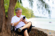 © sutthichai - Asian senior male using mobile phone for video call communication with his family while vacation next to the beach.