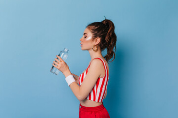 Wall Mural - Young girl with highlighter on face drinks water. Portrait of woman with ponytail on blue background