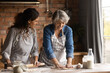 © fizkes - Happy mature grey haired woman with grownup daughter wearing aprons cooking handmade pastry, rolling dough, chatting, having fun, standing in kitchen at home, family spending leisure time together