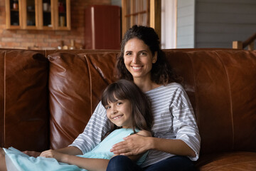 Wall Mural - Head shot portrait smiling mother and little daughter hugging, sitting on couch at home, happy young mum with adorable girl child posing for family photo, looking at camera, enjoying leisure time