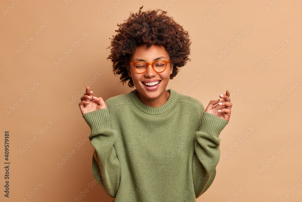 Glad dark skinned woman raises hands has carefree cheerful expression closes eyes smiles toothily wears optical glasses and sweater isolated over beige background. Happiness and joy concept. - obrazy, fototapety, plakaty 