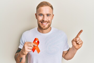 Wall Mural - Young caucasian man holding orange ribbon for leukemia awareness smiling happy pointing with hand and finger to the side