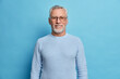 Portrait of handsome bearded European man with grey hair and beard smiles pleasantly looks directly at camera being in good mood has lucky day wears spectacles and sweater isolated over blue wall