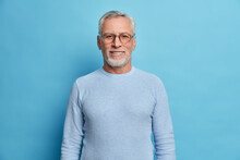 Portrait Of Handsome Bearded European Man With Grey Hair And Beard Smiles Pleasantly Looks Directly At Camera Being In Good Mood Has Lucky Day Wears Spectacles And Sweater Isolated Over Blue Wall