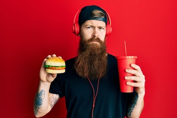 Poster - Redhead man with long beard eating a tasty classic burger and drinking soda skeptic and nervous, frowning upset because of problem. negative person.