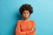 Horizontal shot of dark skinned offended woman with Afro hair keeps arms folded has offensive expression dressed in casual orange jumper being dissatisfied with something expresses negative emotions