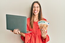 Young Beautiful Woman Holding Laptop And 50 Euros Sticking Tongue Out Happy With Funny Expression.