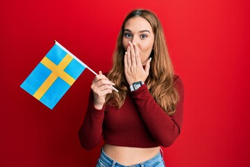 Poster - Young blonde woman holding sweden flag covering mouth with hand, shocked and afraid for mistake. surprised expression