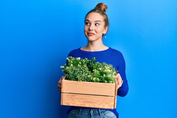 Poster - Young blonde woman holding wooden plant pot smiling looking to the side and staring away thinking.