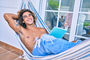 Wall Mural - Young hispanic man relaxed reading book lying on the hammock at terrace.