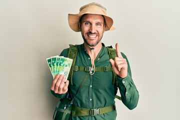 Wall Mural - Handsome man with beard wearing explorer hat holding 50 shekels smiling with an idea or question pointing finger with happy face, number one