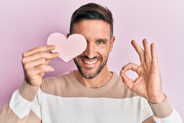 Sticker - Handsome man with beard holding heart shape paper doing ok sign with fingers, smiling friendly gesturing excellent symbol