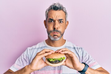 Canvas Print - Middle age grey-haired man eating a tasty classic burger puffing cheeks with funny face. mouth inflated with air, catching air.