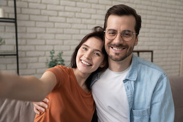 Head shot portrait smiling man wearing glasses and woman hugging, taking selfie, sitting on cozy couch at home, happy couple posing for photo for social network, using gadget, looking at camera