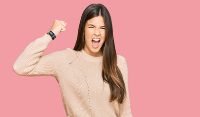 Wall Mural - Young brunette woman wearing casual winter sweater angry and mad raising fist frustrated and furious while shouting with anger. rage and aggressive concept.