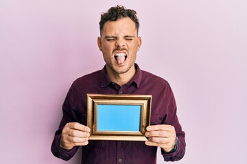 Wall Mural - Young hispanic man holding empty frame sticking tongue out happy with funny expression.