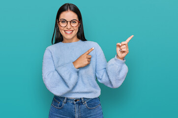 young hispanic woman wearing casual clothes and glasses smiling and looking at the camera pointing w