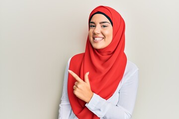 Wall Mural - Young beautiful hispanic girl wearing traditional islamic hijab scarf cheerful with a smile of face pointing with hand and finger up to the side with happy and natural expression on face