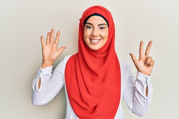 Wall Mural - Young beautiful hispanic girl wearing traditional islamic hijab scarf showing and pointing up with fingers number eight while smiling confident and happy.